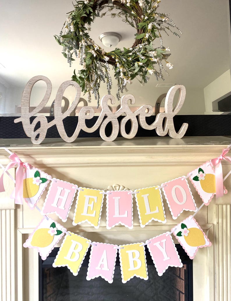 May include: A decorative mantel arrangement featuring a wooden "Blessed" sign, a floral wreath, and two banners. The banners read "HELLO BABY" with lemon and flower accents, perfect for a baby shower or nursery decor.