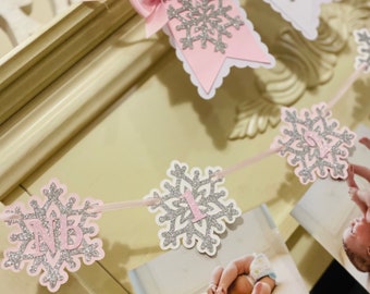 May include: A pink and silver snowflake banner with the letters "NB" and the number "1". The banner is decorated with a pink bow and includes photos of a baby. The banner is likely for a baby's first birthday or a baby shower.