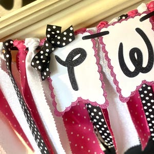 May include: A decorative banner with white, pink, and black polka dot ribbons. The banner features white scalloped-edge pennants with the letters "P" and "W" in black glitter. A black and white polka dot bow adds a festive touch.