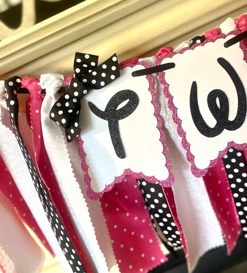 May include: A decorative banner with white, pink, and black polka dot ribbons. The banner features white scalloped-edge pennants with the letters "P" and "W" in black glitter. A black and white polka dot bow adds a festive touch.