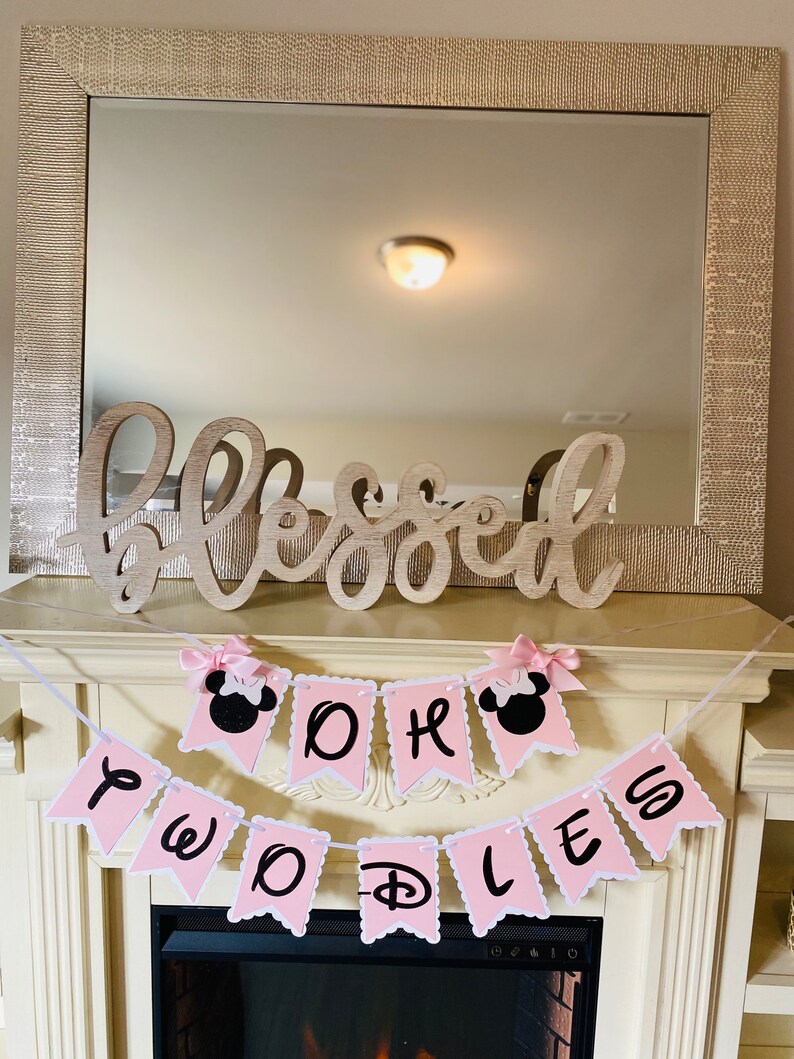 May include: Pink and black banner with the words "Oh Boy Two Doodles" and Mickey Mouse ears on each end. The banner is hanging above a fireplace with a wooden sign that says "Blessed" above it.