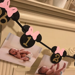 May include: A black and gold glitter banner with Minnie Mouse ears and pink bows, displaying the numbers 0, 1, 2, and 3. The banner includes photos of a baby. A festive decoration for a birthday.