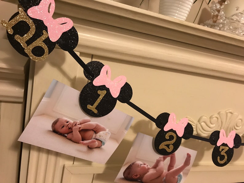 May include: A black and gold glitter banner with Minnie Mouse ears and pink bows, displaying the numbers 0, 1, 2, and 3. The banner includes photos of a baby. A festive decoration for a birthday.