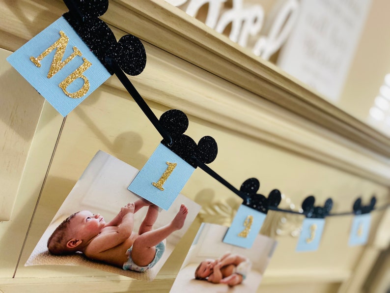 Pode incluir: Uma faixa azul e preta com orelhas do Mickey Mouse e n&uacute;meros de glitter dourado. A faixa diz "NB" e "1". H&aacute; fotos de um beb&ecirc; na faixa.