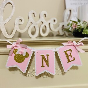 Puede incluir: Bandera rosa y dorada con la palabra "ONE" en letras doradas de purpurina. La bandera tiene un borde festoneado y un lazo rosa en cada extremo. La primera bandera tiene una cabeza de Minnie Mouse dorada de purpurina con un lazo rosa.