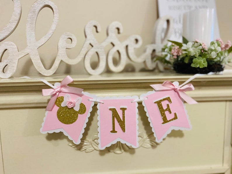 Puede incluir: Bandera rosa y dorada con la palabra "ONE" en letras doradas de purpurina. La bandera tiene un borde festoneado y un lazo rosa en cada extremo. La primera bandera tiene una cabeza de Minnie Mouse dorada de purpurina con un lazo rosa.
