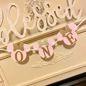 Op de afbeelding: Roze en gouden glitter Minnie Mouse-thema banner met de letters "ONE" op elk van de drie cirkels. De banner hangt aan een wit touw.