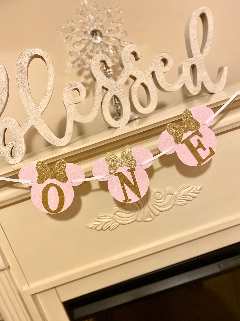 Op de afbeelding: Roze en gouden glitter Minnie Mouse-thema banner met de letters "ONE" op elk van de drie cirkels. De banner hangt aan een wit touw.