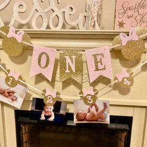 Puede incluir: Pancarta de brillo rosa y dorado con la palabra "ONE" y orejas de Mickey Mouse con los n&uacute;meros del 1 al 4. La pancarta est&aacute; decorada con brillo rosa y dorado. La pancarta est&aacute; colgada en una repisa de chimenea blanca.