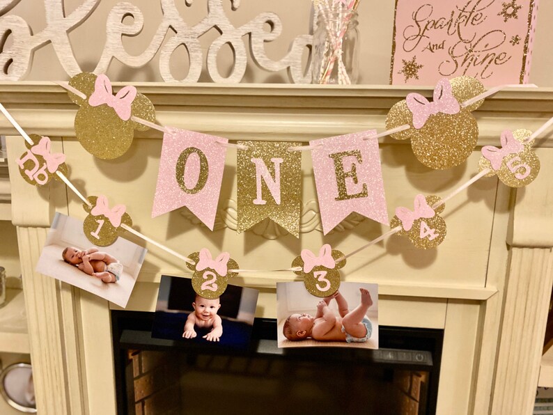 Puede incluir: Pancarta de brillo rosa y dorado con la palabra "ONE" y orejas de Mickey Mouse con los n&uacute;meros del 1 al 4. La pancarta est&aacute; decorada con brillo rosa y dorado. La pancarta est&aacute; colgada en una repisa de chimenea blanca.