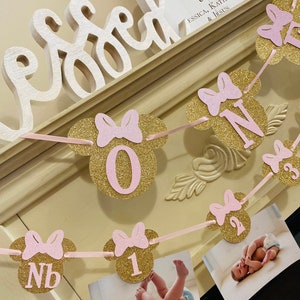 May include: A pink and gold glitter Minnie Mouse themed banner with the numbers 1, 2, 3, and NB. The banner is strung with pink ribbon and has photos of a baby girl in each section. The banner says "Proverbs 31:28, Essica, Kathleen & Jesus".