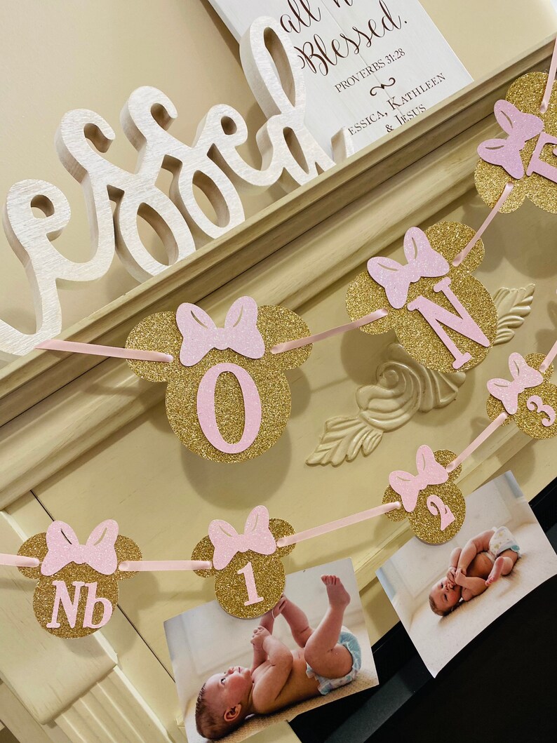 May include: A pink and gold glitter Minnie Mouse themed banner with the numbers 1, 2, 3, and NB. The banner is strung with pink ribbon and has photos of a baby girl in each section. The banner says "Proverbs 31:28, Essica, Kathleen & Jesus".