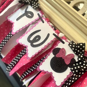 May include: A handmade banner with white, pink, and black polka dot ribbons. The banner spells out "ONE" with glittery black letters on white scalloped flags. Minnie Mouse head with a pink glitter bow.