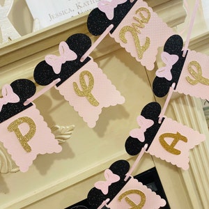 May include: Pink and black banner with gold glitter letters spelling "Happy 2nd Birthday". Each letter is on a pink scalloped flag with a black Minnie Mouse head with a pink bow on top.