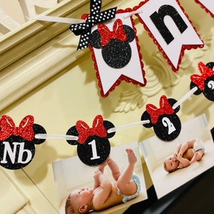 May include: A black and red Minnie Mouse themed banner with the word "one" and numbers 1 and 2. The banner has photos of a baby in a white onesie.