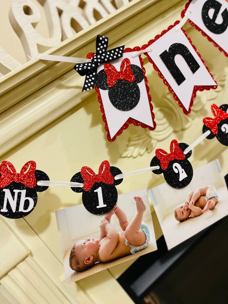 May include: A black and red Minnie Mouse themed banner with the word "one" and numbers 1 and 2. The banner has photos of a baby in a white onesie.