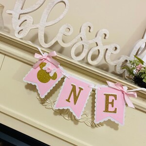 Puede incluir: Bandera rosa y blanca con letras doradas de purpurina que deletrean "ONE" y una cabeza de Minnie Mouse rosa y dorada de purpurina en la primera bandera. La bandera est&aacute; decorada con lazos de cinta de sat&eacute;n rosa.