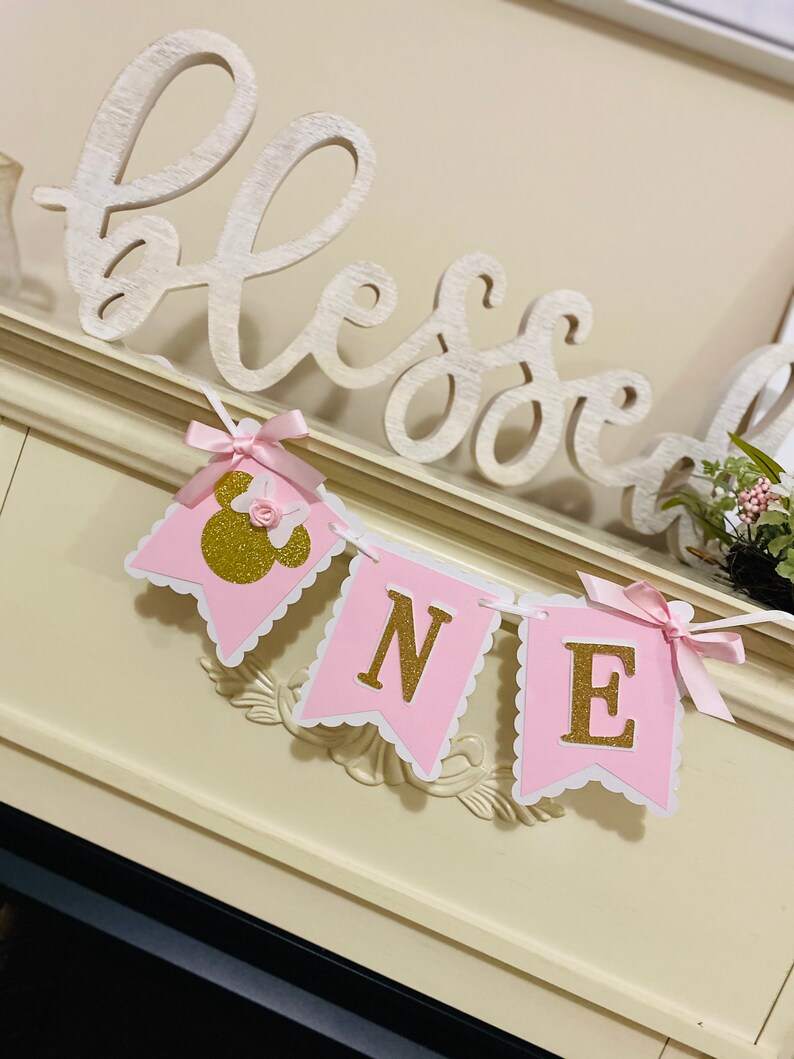 Puede incluir: Bandera rosa y blanca con letras doradas de purpurina que deletrean "ONE" y una cabeza de Minnie Mouse rosa y dorada de purpurina en la primera bandera. La bandera est&aacute; decorada con lazos de cinta de sat&eacute;n rosa.