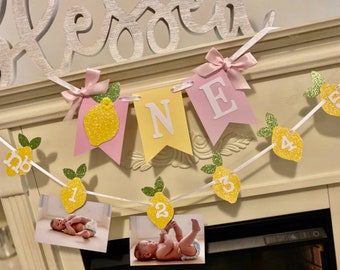 May include: A yellow and pink lemon-themed banner with the word "ONE" in white letters. The banner is decorated with glitter and has small lemon-shaped cutouts with numbers 1 through 6. There are also photos of a baby in the banner.