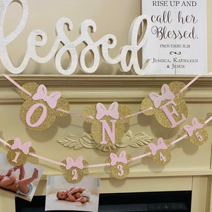May include: A pink and gold glitter banner with the word "ONE" in pink letters and pink and gold glitter Mickey Mouse ears. The banner is strung with pink ribbon and has photos of a baby girl in each of the Mickey Mouse ears. The banner also includes the text "Rise up and call her blessed. Proverbs 31:28 Jessica, Kathleen & Jesus."