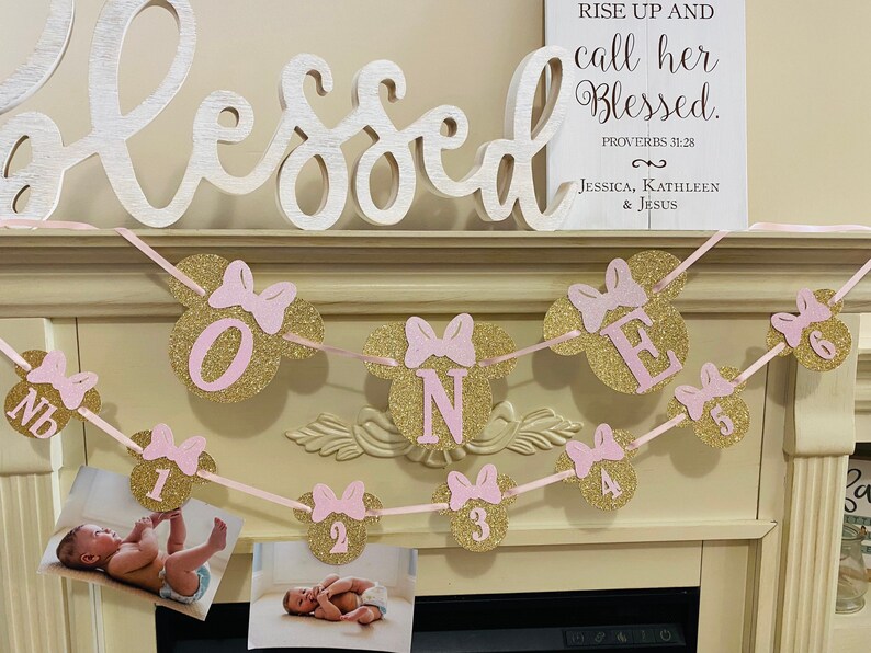 May include: A pink and gold glitter banner with the word "ONE" in pink letters and pink and gold glitter Mickey Mouse ears. The banner is strung with pink ribbon and has photos of a baby girl in each of the Mickey Mouse ears. The banner also includes the text "Rise up and call her blessed. Proverbs 31:28 Jessica, Kathleen & Jesus."