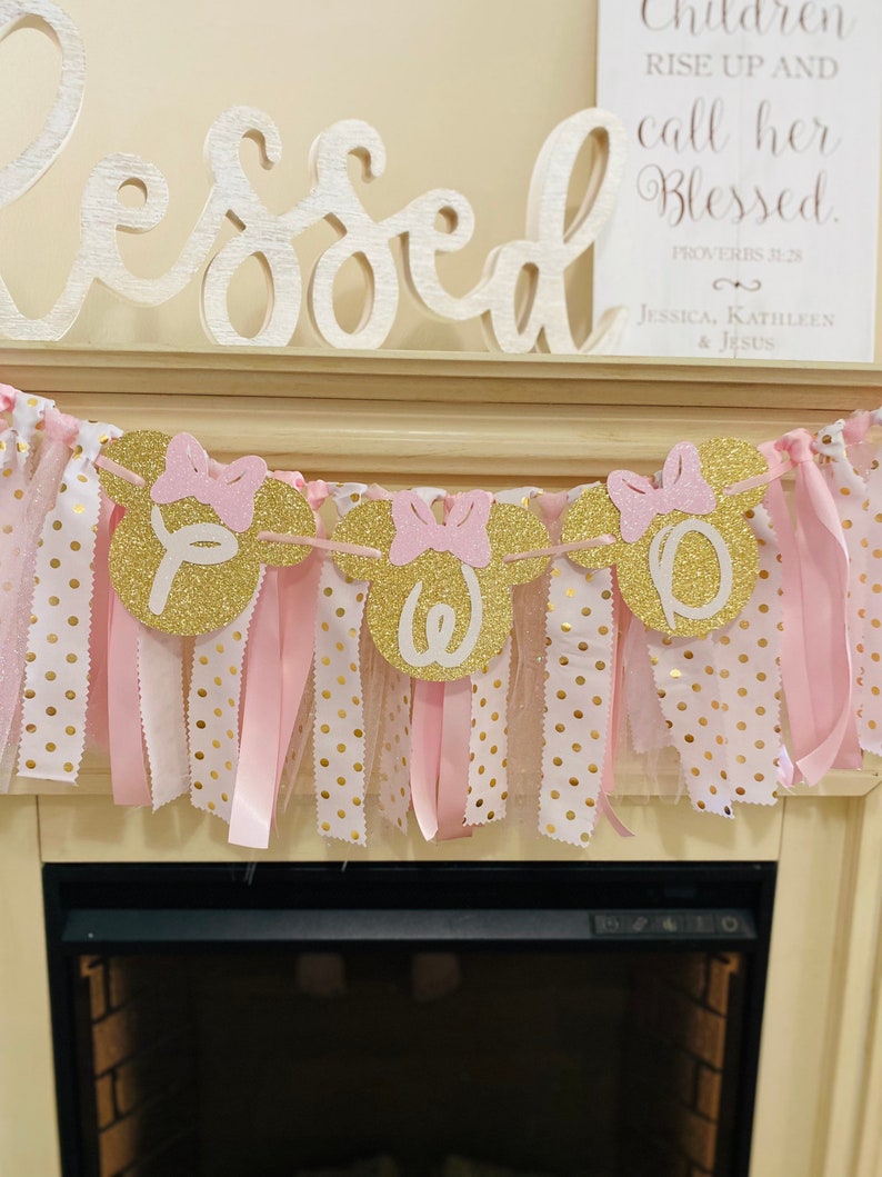 May include: A decorative banner with gold glitter mouse head cutouts spelling "OWE" with pink bows. The banner is adorned with pink and white polka dot ribbons and tulle, creating a festive and celebratory look.