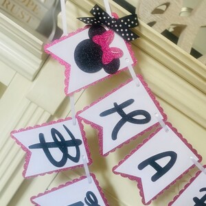 May include: A pink and white banner with black letters that reads "Happy 1st Birthday". The banner has a pink glitter border and a black and white polka dot bow with a pink bow on top.