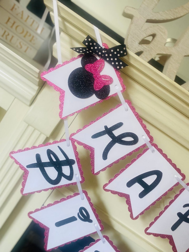 May include: A pink and white banner with black letters that reads "Happy 1st Birthday". The banner has a pink glitter border and a black and white polka dot bow with a pink bow on top.