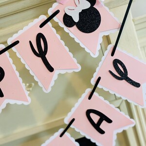 Puede incluir: Bandera rosa y negra con las palabras "Feliz cumplea&ntilde;os" en letras negras. La bandera presenta una cabeza de Minnie Mouse de purpurina negra y blanca con un lazo rosa.