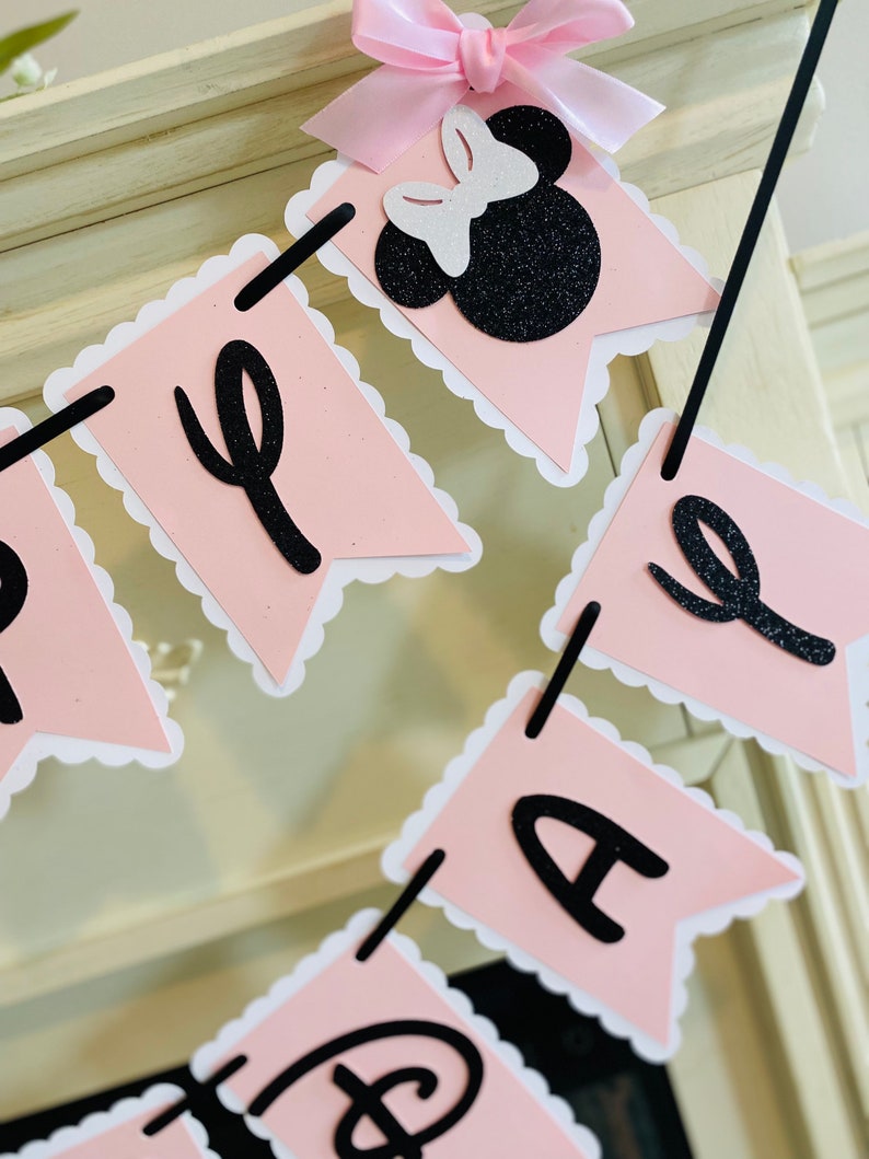 Puede incluir: Bandera rosa y negra con las palabras "Feliz cumplea&ntilde;os" en letras negras. La bandera presenta una cabeza de Minnie Mouse de purpurina negra y blanca con un lazo rosa.