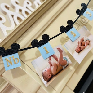 Pode incluir: Uma faixa azul e dourada com orelhas de Mickey Mouse pretas, apresentando fotos de um beb&ecirc; em diferentes idades. A faixa inclui o texto "NB" e "1".