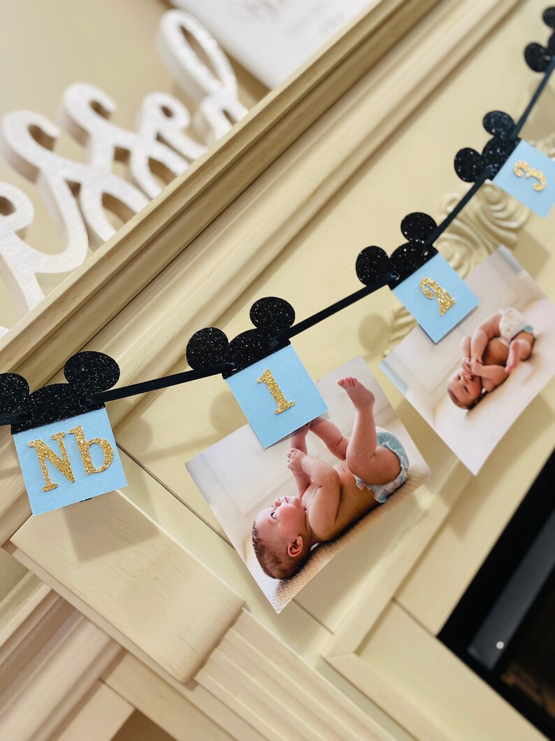 Pode incluir: Uma faixa azul e dourada com orelhas de Mickey Mouse pretas, apresentando fotos de um beb&ecirc; em diferentes idades. A faixa inclui o texto "NB" e "1".