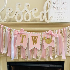 May include: A pink and gold "Two" banner with a Minnie Mouse silhouette, ribbons, and bows. The banner is decorated with gold glitter letters and is hanging above a fireplace. The word "Blessed" is displayed above the banner.