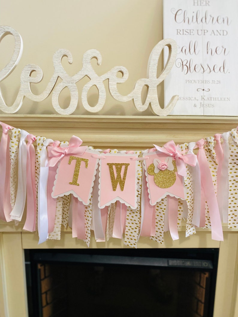 May include: A pink and gold "Two" banner with a Minnie Mouse silhouette, ribbons, and bows. The banner is decorated with gold glitter letters and is hanging above a fireplace. The word "Blessed" is displayed above the banner.