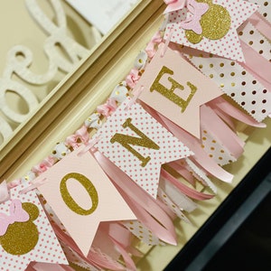 May include: A pink and gold "ONE" banner with Minnie Mouse accents. The banner features pennants with gold glitter letters and polka dot patterns, along with pink ribbons and bows. The banner is designed for a first birthday celebration.