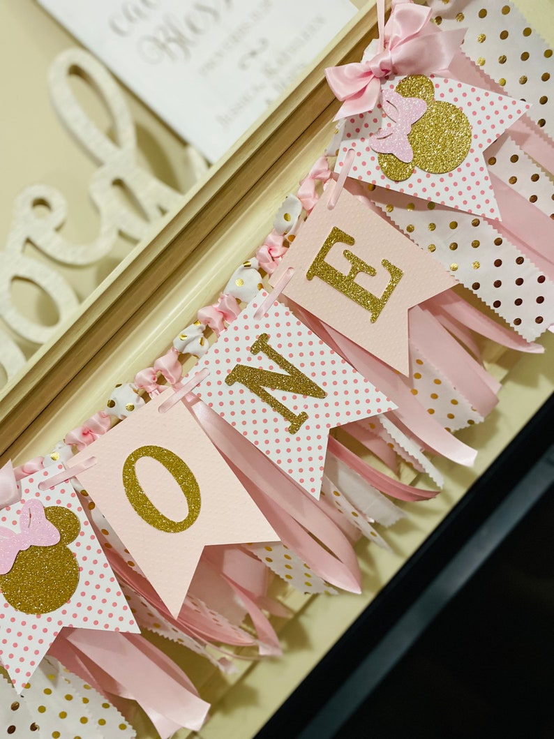 May include: A pink and gold "ONE" banner with Minnie Mouse accents. The banner features pennants with gold glitter letters and polka dot patterns, along with pink ribbons and bows. The banner is designed for a first birthday celebration.