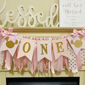 May include: A pink and gold "ONE" banner with Minnie Mouse accents. The banner has pink ribbons and gold polka dots. The word "Blessed" is displayed above the banner, and a framed sign reads "Her Children Rise Up and Call Her Blessed."