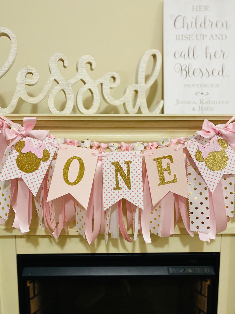 May include: A pink and gold "ONE" banner with Minnie Mouse accents. The banner has pink ribbons and gold polka dots. The word "Blessed" is displayed above the banner, and a framed sign reads "Her Children Rise Up and Call Her Blessed."