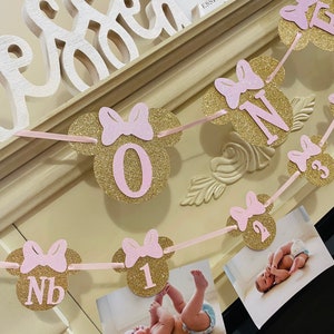 May include: A pink and gold glitter Minnie Mouse themed banner with the numbers 1, 2, and 3. The banner is strung with pink ribbon and has pink bows on each of the Minnie Mouse ears. The banner is hanging on a white mantle with a sign that says "Blessed" and "Proverbs 3:28".