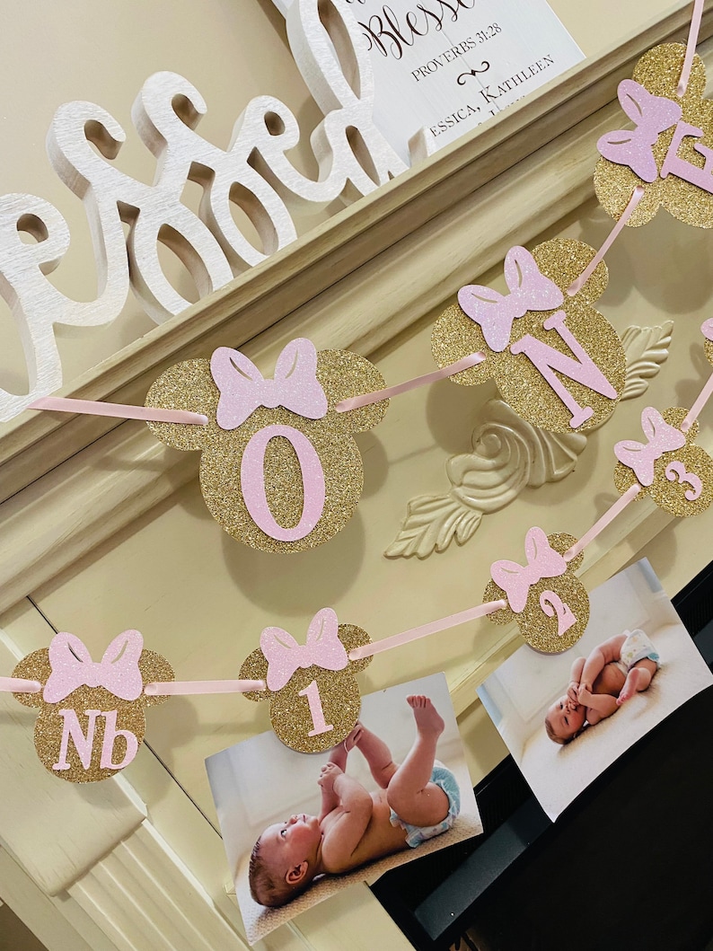 May include: A pink and gold glitter Minnie Mouse themed banner with the numbers 1, 2, and 3. The banner is strung with pink ribbon and has pink bows on each of the Minnie Mouse ears. The banner is hanging on a white mantle with a sign that says "Blessed" and "Proverbs 3:28".