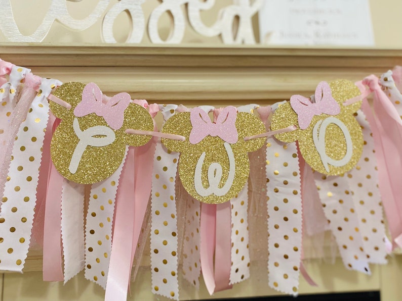May include: A festive banner with gold glitter Mickey Mouse head shapes, each with a pink bow and a white letter spelling "One". The banner is adorned with pink and white polka dot ribbons, perfect for a birthday celebration.