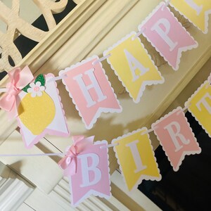 May include: A pink and yellow banner with the words "Happy Birthday" in white letters. The banner is decorated with a lemon and a pink bow.