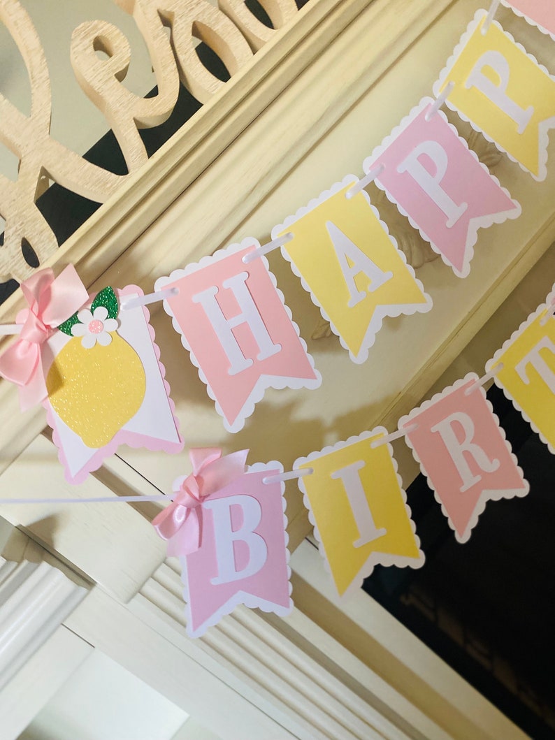 May include: A pink and yellow banner with the words "Happy Birthday" in white letters. The banner is decorated with a lemon and a pink bow.
