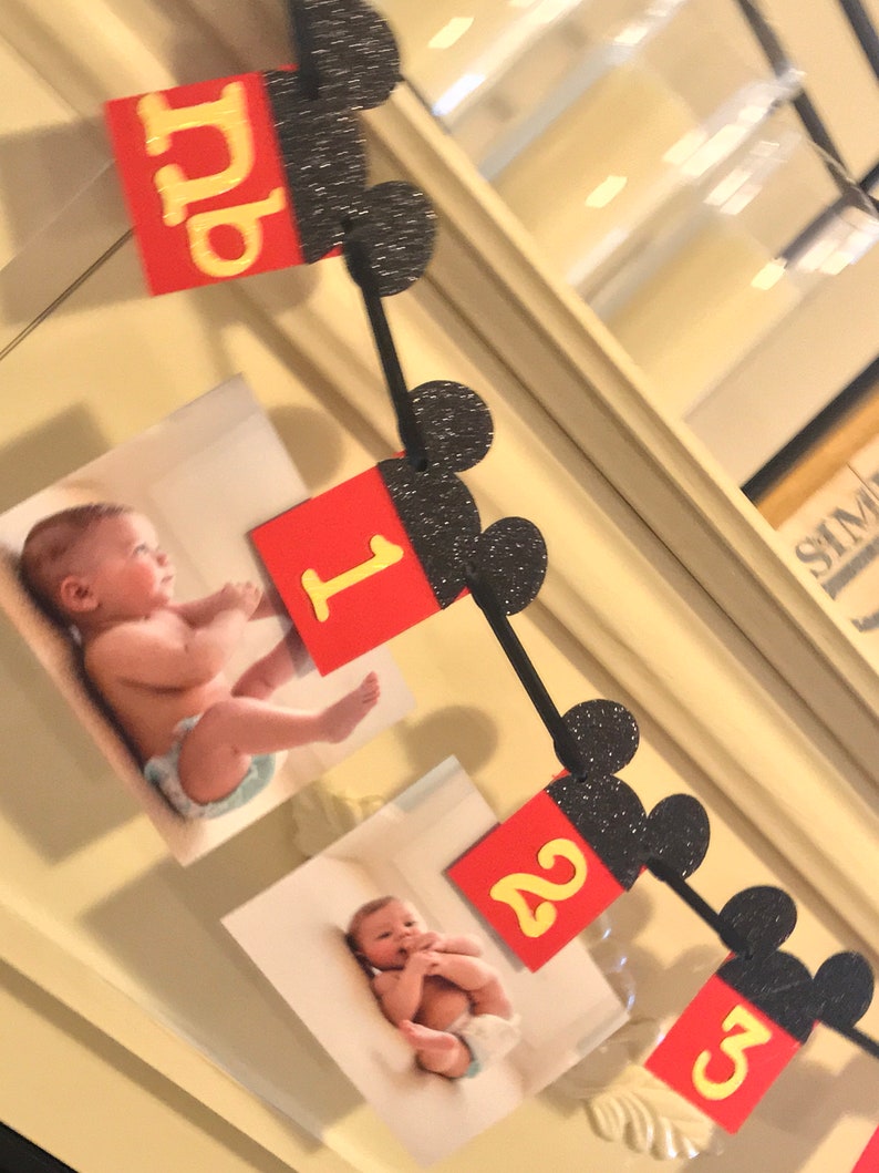 May include: A black and red Mickey Mouse themed banner with pictures of a baby and the numbers 1, 2, and 3. The banner is made of felt and has glitter on the Mickey Mouse ears.