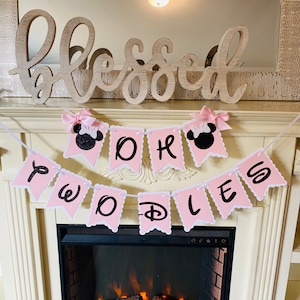 May include: Pink and black banner with the words "Oh the Places You'll Go" and a Mickey Mouse head on each end. The banner is hanging above a fireplace with a wooden sign that says "blessed" above it.