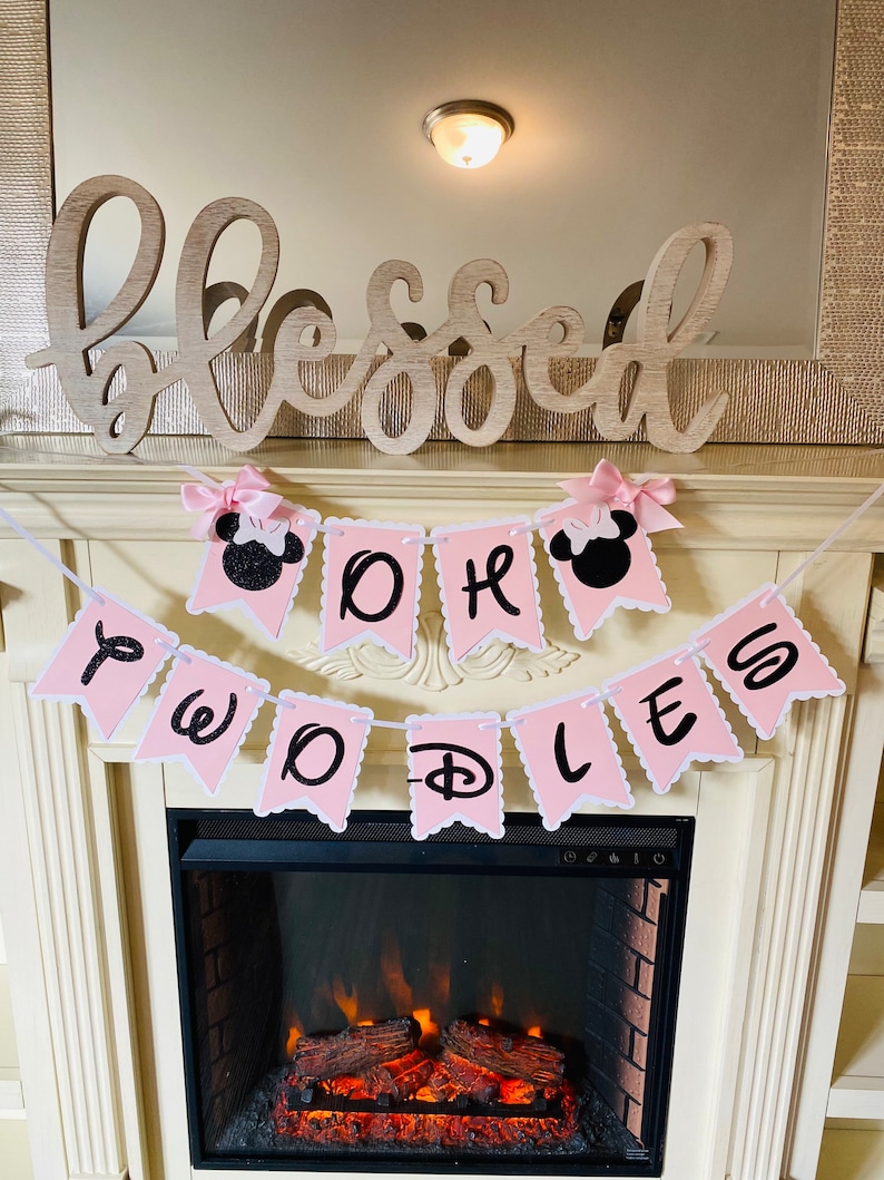 May include: Pink and black banner with the words "Oh the Places You'll Go" and a Mickey Mouse head on each end. The banner is hanging above a fireplace with a wooden sign that says "blessed" above it.