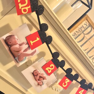 May include: A Mickey Mouse themed baby milestone banner with black glitter ears and red and yellow numbers. The banner includes photos of a baby at different ages.