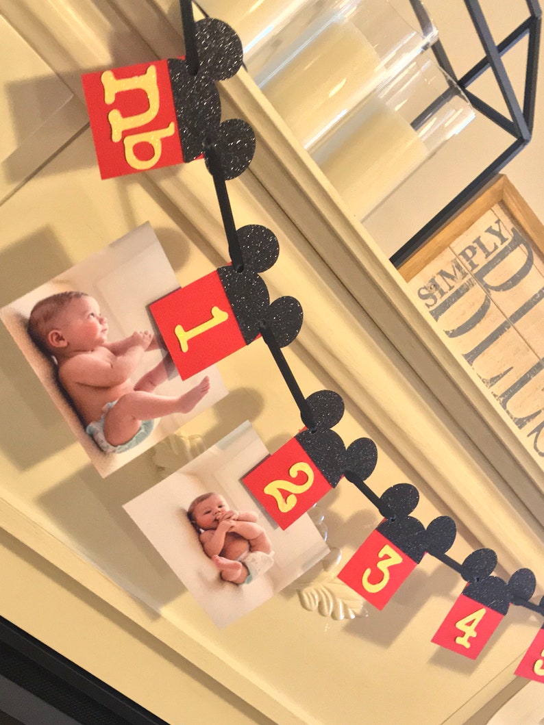 May include: A Mickey Mouse themed baby milestone banner with black glitter ears and red and yellow numbers. The banner includes photos of a baby at different ages.