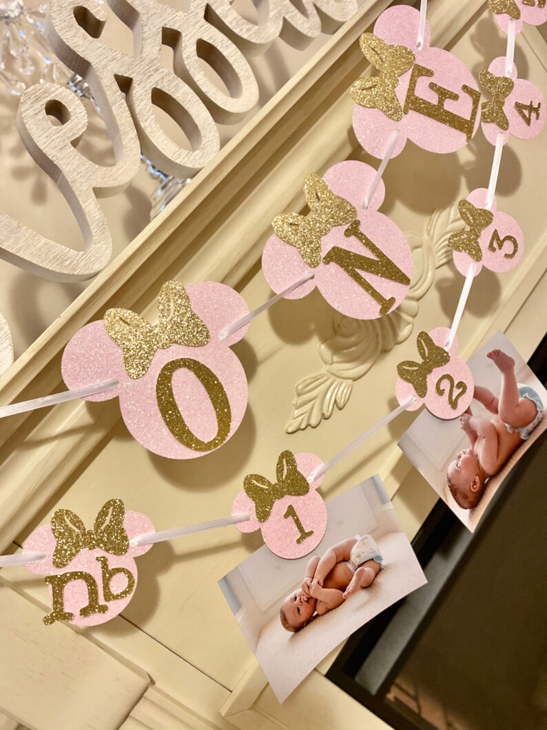 May include: Pink and gold glitter Minnie Mouse themed banner with numbers 1 through 4 and the letter "M" for a first birthday party. Each number is on a separate pink circle with a gold glitter bow. The banner is strung with white ribbon.