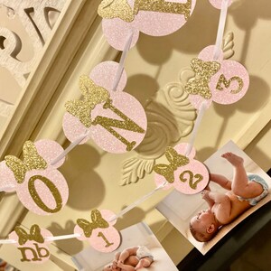 May include: Pink and gold glitter Minnie Mouse themed banner with the numbers 0-3 and the letters "nb" for a first birthday party.